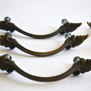 May include: Set of antique bronze-toned drawer pulls. Each pull features a curved design with ornate, leaf-like detailing and a clear, round knob. These decorative pulls are ideal for furniture restoration projects.