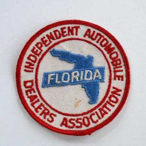 Könnte beinhalten: Ein gestickter Aufnäher in Rot, Weiß und Blau mit dem Text "Independent Automobile Dealers Association" und "Florida" auf einem weißen Banner. Der Aufnäher zeigt eine Umrandung des Bundesstaates Florida in Blau.