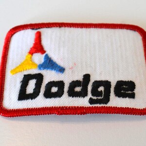 Op de afbeelding: Geborduurde patch met een witte achtergrond, rode rand en het woord "Dodge" in zwart. De patch heeft een kleurrijk logo met rode, gele en blauwe vormen. Deze vintage patch is waarschijnlijk afkomstig uit de auto-industrie.