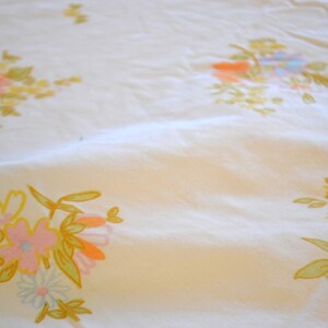 Könnte beinhalten: Cremefarbener Stoff mit einem sich wiederholenden Blumenmuster. Die Blumen sind in Rosa-, Gelb-, Orange- und Blautönen gehalten, mit grünen Blättern. Der Stoff wirkt leicht zerknittert.