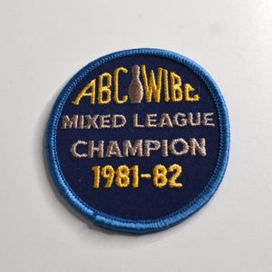 May include: A circular embroidered patch with a dark blue background and a light blue border. It features yellow text reading ABC WIBC, the words MIXED LEAGUE CHAMPION in silver, and the year 1981-82 in yellow, with a small bowling pin icon.