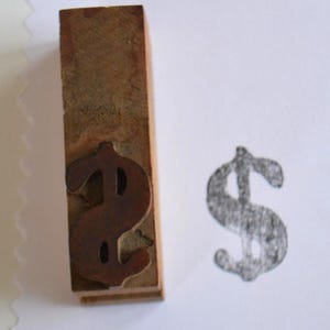 Könnte beinhalten: Ein Holzstempel mit einem Dollarzeichen, neben einem gedruckten Dollarzeichen-Bild. Der Stempel ist braun, mit einem dunkleren braunen Dollarzeichen. Das gestempelte Bild ist in Schwarz.