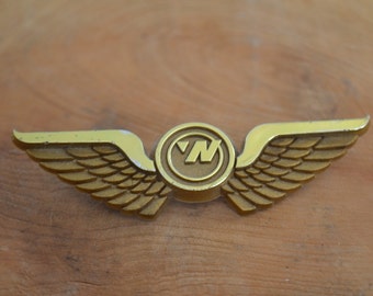 Puede incluir: Un pin de metal dorado con una letra "N" estilizada en un círculo y alas a cada lado.