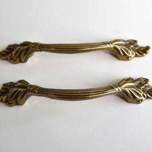 May include: Two matching brass drawer pulls featuring a central fluted bar design that transitions into detailed leaf-like motifs at each end. The metal hardware has a warm, aged finish with visible grooves and a curved, elongated shape.