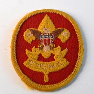 May include: A vintage Boy Scouts of America patch. The patch is circular with a red background and a yellow embroidered border. The central design features a fleur-de-lis with an eagle and a banner with text.