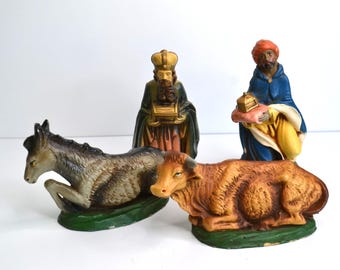 May include: A vintage Nativity scene set with four figurines. It includes a donkey, a cow, and two figures of the Three Wise Men. The figures are painted in various colours, including brown, gold, blue, and green.