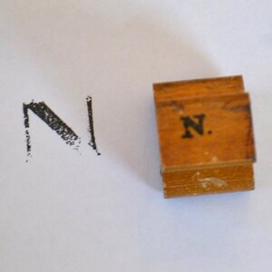 Könnte beinhalten: Ein Holzbuchstaben-Stempel mit dem Buchstaben "N" darauf gedruckt. Der Stempel ist braun und hat eine leicht abgenutzte Optik. Der Buchstabe "N" ist in Schwarz gedruckt.