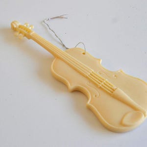 May include: A light beige violin-shaped ornament with a wire loop for hanging. The instrument's details, including the strings and scroll, are molded into the plastic. A simple, decorative item.