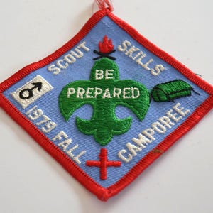 May include: A vintage Scout Camporee patch with a light blue background and red border. The patch features the words "SCOUT SKILLS CAMPORÉE" and "1979 FALL" in white, along with a green fleur-de-lis and the words "BE PREPARED".