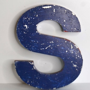 May include: A capital letter S made of wood with a distressed finish. The surface is painted in a deep blue color with visible white paint chips and scratches throughout, revealing the texture of the material underneath the blue paint layer.