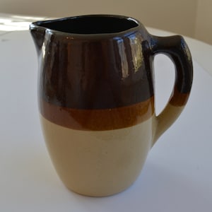 May include: A brown, tan, and cream colored ceramic pitcher with a handle. The pitcher has a glazed finish.