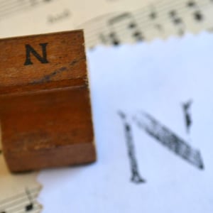 May include: A vintage wooden stamp with the letter "N" printed in black ink. The stamp is resting on a sheet of paper with a printed "N" and musical notes in the background. The stamp has a rectangular shape.
