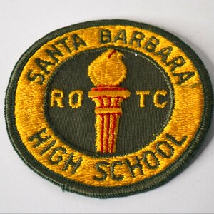 May include: A circular embroidered patch with a dark olive green centre and a gold border. The text SANTA BARBARA HIGH SCHOOL is stitched in gold along the border. A gold torch icon sits in the centre with the letters RO and TC on either side.