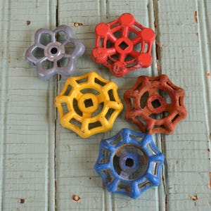 May include: Five vintage water valve handles in various colors: gray, red, yellow, rust, and blue. Each handle has a unique starburst design with a square center hole. The handles are arranged on a weathered, light blue wooden surface.