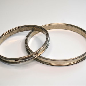 May include: Two silver-toned metal hoops, one slightly smaller than the other, are displayed against a white background. The smaller hoop has a spring mechanism. The hoops appear to be vintage or antique, with a slightly worn appearance.