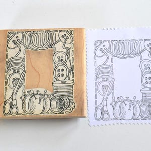 May include: A wooden stamp with a rectangular design featuring sewing tools and notions. The design includes spools of thread, buttons, scissors, and pincushions. A stamped image of the design is next to the stamp.