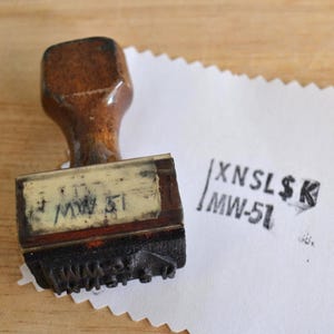 May include: A brown wooden rubber stamp with a rectangular handle and a label reading MW-51. The stamp is positioned next to a white paper with a black ink impression that reads XNSL$K and MW-51, featuring a vertical line to the left of the text.