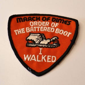 Op de afbeelding: Een oranje viltpatch met zwarte stiksels, met de tekst "March of Dimes Order of the Battered Boot I Walked" in witte borduurwerk. In het midden is een wit geborduurd beeld van een versleten laars.