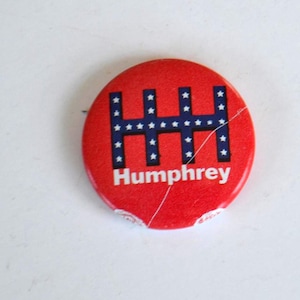 May include: A red circular button with a blue and white design and the name "Humphrey" in white text. The blue design features a series of connected vertical and horizontal lines with white stars. The button has a crack.