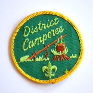 Puede incluir: Parche bordado redondo, verde con borde amarillo. Presenta las palabras "District Camporee" en verde, una escena de fogata y un símbolo de flor de lis. Ideal para actividades de campamento.