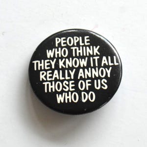 Può includere: Una spilla rotonda nera con testo bianco che recita: PEOPLE WHO THINK THEY KNOW IT ALL REALLY ANNOY THOSE OF US WHO DO. La spilla ha una finitura liscia e lucida e un design semplice e audace con caratteri sans-serif chiari e leggibili.