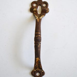Puede incluir: Una cubierta de ojo de cerradura antigua y ornamentada con un acabado marrón oxidado y desgastado. La pieza metálica presenta un diseño floral decorativo en la parte superior e inferior, con un mango largo y delgado. Para muebles antiguos.