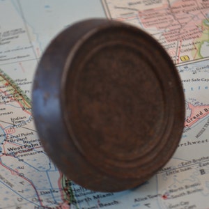 May include: A close-up of a round, brown, metal lid with a slightly raised rim. The lid has a worn, aged appearance.