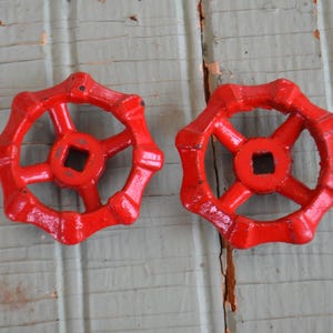 May include: Two red, cast iron valve handles with a starburst design. Each handle has a square center and a series of rounded spokes. The handles are set against a weathered, light-colored wooden surface.