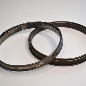 Peut inclure: Deux cerceaux métalliques gris foncé, peut-être pour un tambour ou un autre objet circulaire. Les cerceaux ont une apparence légèrement vieillie, suggérant l'âge et l'utilisation. Ils sont posés sur une surface blanche.