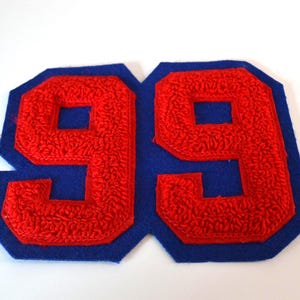 May include: A red and blue felt patch featuring the number "99". The number is in a bold, textured font, stitched in red, and set against a blue background. The patch is likely for a sports uniform or varsity jacket.