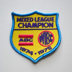 May include: A yellow shield-shaped patch with blue trim. The patch says "Mixed League Champion" in purple lettering. It also has the letters "ABC" in red and a circle with "WIBC" in purple lettering. The years "1974-1975" are printed below the lettering.