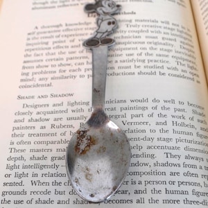 Puede incluir: Una cuchara de plata vintage con una figura de Mickey Mouse en la parte superior, apoyada en un libro abierto. La cuchara muestra signos de desgaste y edad. Las páginas del libro están amarillentas, con texto visible.