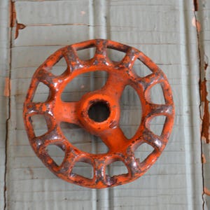 May include: An orange, circular valve wheel with a series of open spokes. The wheel has a central hub with a hole and shows signs of rust and wear. The background is a weathered, light blue surface.