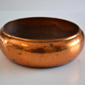 Puede incluir: Un cuenco redondo y poco profundo hecho de cobre pulido con un acabado metálico de color naranja parduzco. La superficie exterior muestra signos visibles de desgaste, incluyendo pequeñas manchas oscuras e imperfecciones en el cuerpo.