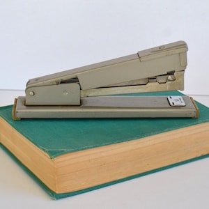 May include: A silver stapler with a black handle sits on top of a green and brown book. The stapler has a metal base and a lever on the top.