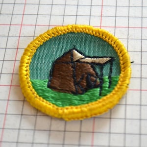 Könnte beinhalten: Runder, gestickter Aufnäher mit gelbem Rand, der ein braunes Zelt mit weißem Dach auf grünem und blaugrünem Hintergrund darstellt. Das Design deutet auf ein Camping- oder Outdoor-Thema hin.