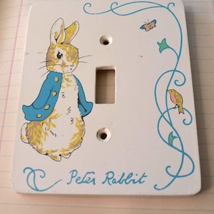 May include: A white light switch cover featuring a painted illustration of Peter Rabbit in a blue jacket. The cover has a rectangular opening for the switch and two screw holes. The words "Peter Rabbit" are written in blue cursive.