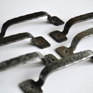 May include: Set of six rustic, hammered metal handles. The handles are dark gray with a textured surface and a curved design. Each handle has a rectangular base with two screw holes for mounting.