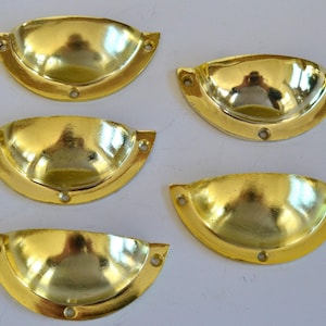 Puede incluir: Cinco tiradores de armario en forma de copa de color dorado con un acabado liso y brillante. Los tiradores están dispuestos en semicírculo sobre una superficie blanca.