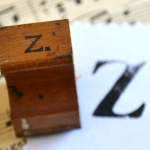 Könnte beinhalten: Ein Holzstempel mit dem Buchstaben "Z" auf der Oberseite. Der braune Stempel weist Gebrauchsspuren auf. Ein gedrucktes "Z" in schwarzer Tinte ist auf weißem Papier zu sehen, was auf seine Verwendung zum Drucken hindeutet.