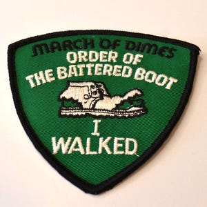 Op de afbeelding: Groen geborduurd patch met witte tekst die "March of Dimes Order of the Battered Boot I Walked" luidt. Een witte cartoon-laars bevindt zich in het midden van het patch.