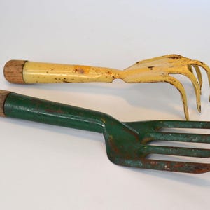 May include: Two garden hand tools: a yellow rake and a green cultivator. The rake has curved tines and a wooden handle. The cultivator has three tines and a wooden handle. Both tools show signs of wear.