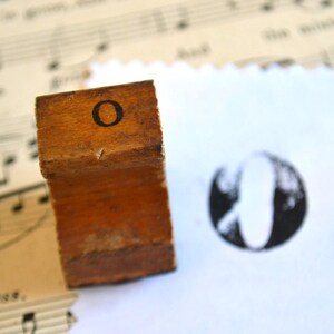 Könnte beinhalten: Ein Holzstempel mit dem Buchstaben "O" und einem schwarzen Tintenabdruck auf weißem Papier. Der Stempel ist rechteckig und hat den Buchstaben "O" auf der Oberseite aufgedruckt. Der Stempel ist braun und weist Gebrauchsspuren auf.