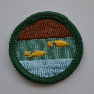 Può includere: Una toppa ricamata circolare con un bordo verde scuro. Il design presenta una sezione superiore marrone, un'area centrale verde con due forme gialle ricamate e una sezione inferiore azzurra che rappresenta l'acqua con cuciture testurizzate.