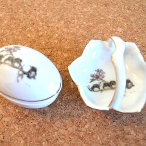 Może przedstawiać: Białe, porcelanowe pudełko na bibeloty w kształcie jajka i mała miseczka z uchwytem. Oba elementy zdobione wzorem kwiatowym i ptasim w odcieniach różu, zieleni i czerni, ze złotym obramowaniem. Miseczka ma falisty brzeg.