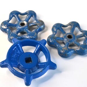 May include: Three vintage blue metal valve handles with a flower-like design. The handles have a distressed, aged appearance with visible wear and tear. The central hole is square-shaped.