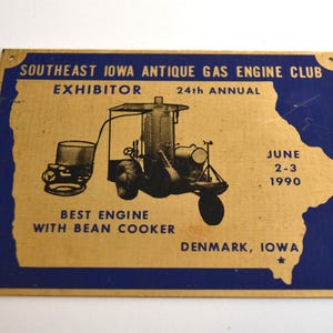 Può includere: Una targa rettangolare in metallo con bordo blu e centro color oro che riporta il testo SOUTHEAST IOWA ANTIQUE GAS ENGINE CLUB, EXHIBITOR 24th ANNUAL, JUNE 2-3 1990, BEST ENGINE WITH BEAN COOKER e DENMARK, IOWA con un grafico di un motore.