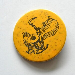 Könnte beinhalten: Ein runder, gelber Ansteckbutton mit gesprenkelter Textur. In der Mitte befindet sich eine schwarze Strichzeichnung eines Saxophons, das mit Noten verflochten ist, sowie das Wort JAZZ in einer stilisierten, handgezeichneten Schrift darunter.