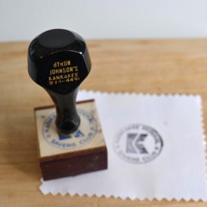 May include: A black wooden rubber stamp with a handle featuring gold text that reads BYRON JOHNSON'S KANKAKEE 933-4491. The square base has a blue ink design, and a white paper with a stamped black circular logo is positioned next to the stamp.