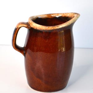 Pode incluir: Um jarro de cerâmica castanho com uma pega e um bico. O jarro tem um acabamento brilhante e uma borda castanha mais clara. O design é simples e clássico, adequado para servir bebidas ou como objeto decorativo.